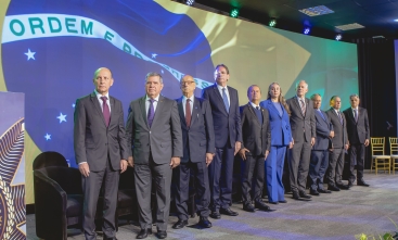 Grupo de autoridades em pé, lado a lado, em frente à imagem projetada da bandeira do Brasil, em evento formal.