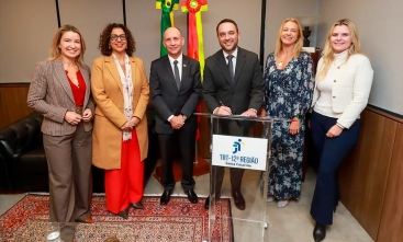 Grupo de seis pessoas posa sorridente ao lado de púlpito do TRT-12 durante cerimônia formal com bandeiras e termo de adesão ao fundo.