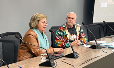 Uma mulher e um homem estão sentados em um palco fazendo uma palestra
