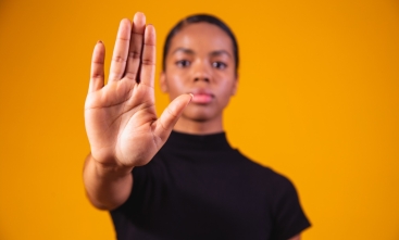 Uma mulher negra está com a mão estendida para frente, com rosto sério e vestida de preto