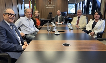 Quatro homens e duas mulheres sentados em torno de uma mesa retangular sorriem para a foto, em uma sala com janelas abertas