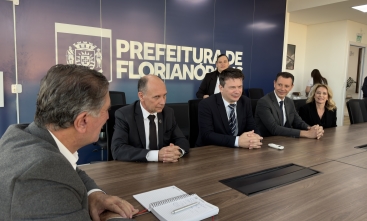 Foto de cinco pessoas sentadas à mesa, sendo quatro homens e uma mulher. Ao fundo, na parede, escrito Prefeitura de Florianópolis