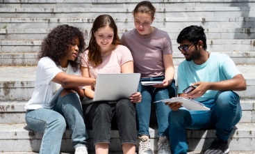 Grupo de quatro estudantes sentados em escadaria ao ar livre, olhando para um notebook com atenção.