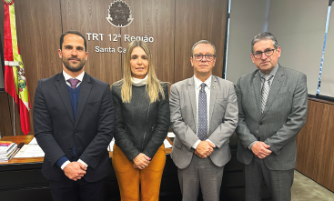 Quatro pessoas posam lado a lado em frente ao letreiro “TRT 12ª Região – Santa Catarina”, dentro de um gabinete. À esquerda, bandeira de Santa Catarina. Todos estão com trajes formais.