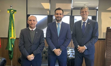 Três homens de terno, lado a lado, posam para foto, dentro de uma sala. À esquerda, aparece a bandeira do Brasil