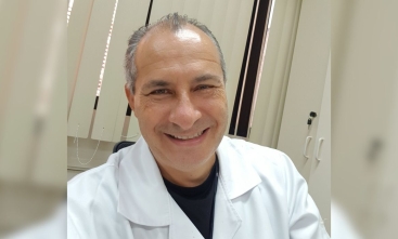 Homem posa para foto sorrindo. Ele está de jaleco em um consultório.