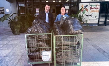 Imagem de dois carrinhos de transporte recheados de sacos pretos com artigos dentro. Um homem e uma mulher sorriem para a foto 