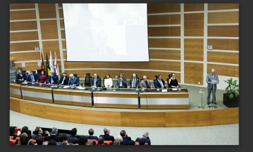 Cerimônia formal em auditório. Autoridades sentadas à mesa no palco, homem discursa no púlpito. Público assiste. Telão ao fundo exibe participante remoto. Bandeiras à esquerda, planta decorativa à direita.