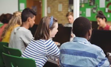 Grupo de alunos, a maioria de costas, sentados em frente a monitores de computador
