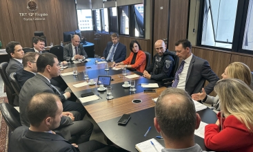 Reunião no TRT-SC com autoridades e representantes, incluindo uma agente da Guarda Municipal, em torno de mesa com anotações e copos d’água.