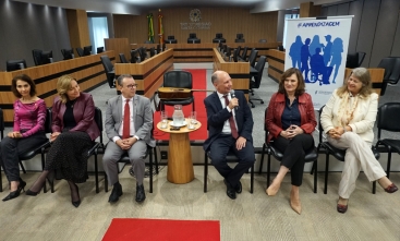 Quatro mulheres e dois homens sentados à frente de um auditório, em cadeiras. Ao fundo, um violão e um banner escrito Aprendizagem