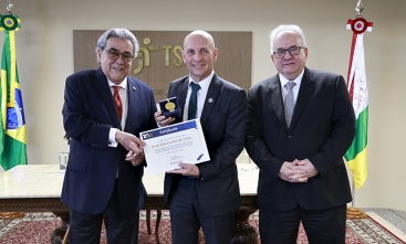 Três homens de terno e gravata estão em pé, posando para a foto. O homem que estão no centro segura um certificado e uma medalha