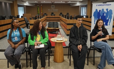 Três jovens e uma mulher madura sentados em cadeiras, dentro de uma ampla sala de julgamentos. Uma das jovens segura o microfone e sorri