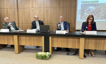 Três homens e uma mulher sentados à mesa de um auditório. Um dos homens fala ao microfone