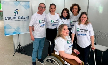 Um homem e quatro mulheres em pé e uma mulher na cadeira de rodas estão posando para a foto. Ao lado deles um banner escrito: Justiça do Trabalho