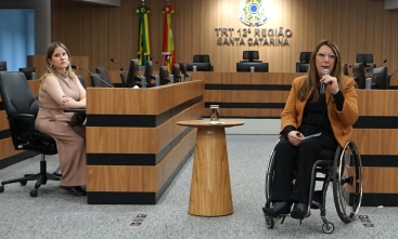Foto de uma mulher de cadeiras de rodas dando uma palestra. No fundo um painel escrito Tribunal Regional do Trabalho da 12ª Região.