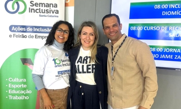 Três pessoas sorriem para foto durante a Semana Inclusiva Santa Catarina, em frente a banner do evento.