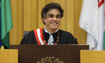 Homem de cerca de 60 anos, vestido com toga de juiz e faixa, sorri 