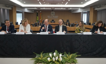Três homens e três mulheres sentadas à mesa de um evento, coberta por uma toalha preta e um buquê de flores no chão.