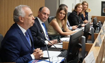 Foto de uma mesa de honra de um evento, com um homem falando e mais homens e mulheres sentados olhando o palestrante