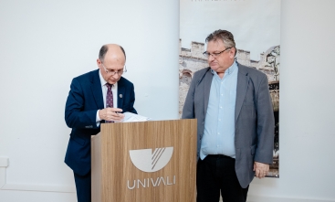 Dois homens estão posicionados ao lado de um púlpito de madeira com o nome e logotipo da UNIVALI. Um deles escreve ou assina um documento sobre o púlpito.