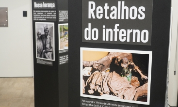 Totem de uma exposição mostrando a foto de um trabalhador no meio de muitos tecidos. O texto acima da foto é "Retalhos do Inferno".