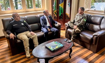 Três homens, dois civis e um militar, conversam em sala com sofás, bandeiras e livro sobre a mesa.