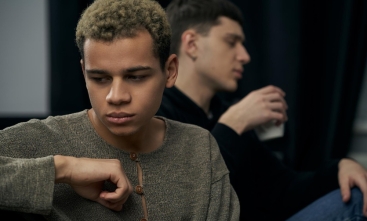 Fotografia ilustrativa de homem jovem pensativo em primeiro plano, outro desfocado ao fundo segurando copo