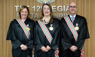 A imagem mostra duas magistradas e um magistrado posando lado a lado, usando togas pretas e faixas nas cores verde, branca e vermelha, com medalhas douradas presas ao peito.