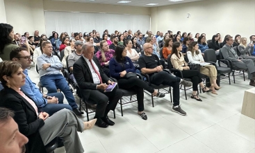 Foto em diagonal mostra cerca de 90 pessoas sentadas em cadeiras dentro de um auditório