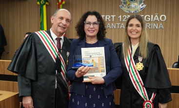 Três pessoas sorridentes posam em solenidade no TRT-12; ao centro, mulher segura troféu e livro comemorativo.