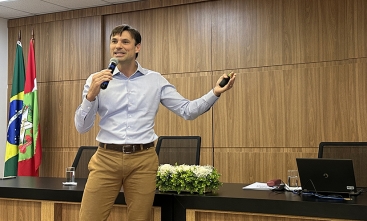 Ao centro, um homem jovem fala ao microfone. Ao fundo dele, um painel de madeira