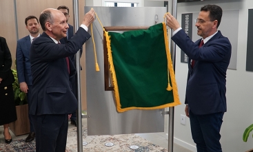 Um homem calvo, de cerca de 60 anos, de terno, e outro homem moreno, também de terno, retirar um pano verde que está cobrindo uma placa de metal