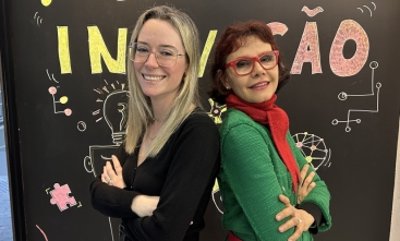 Duas mulheres, uma de costas para a outra, posam de braços cruzados