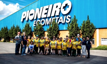Foto de um grupo de pessoas, algumas com colete amarelo, em pé, em frente a uma empresa com o nome Pioneiro Ecometais