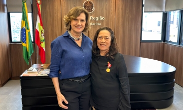 Duas mulheres, uma loira e outra morena, posam sorridentes para a foto. A morena tem uma medalha fixada na roupa