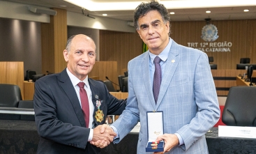 Dois homens de terno, com cerca de 60 anos, se cuprimentam. Um deles, de terno claro, exibe uma caixa contendo uma medalha.