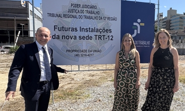 Um homem de terno e duas mulheres ao lado de uma placa de cerca de 2 metros de altura sobre um terreno vazio