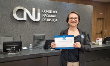 Mulher sorridente segurando o certificado. Ao fundo a logo do CNJ