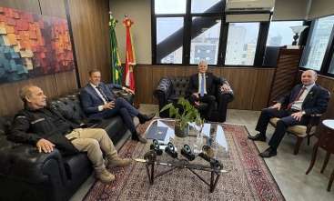 Três homens de terno e um com uniforme de segurança sentados em poltronas e cadeiras em uma sala arejada