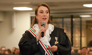 Mulher de toga e faixa fala ao microfone