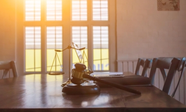 Foto de um martelo da justiça em cima de uma mesa de madeira. Ao fundo uma janela com o sol batendo como reflexo.