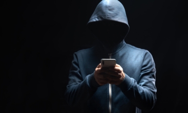 Pessoa de moletom com capuz, rosto oculto, usando celular em ambiente escuro