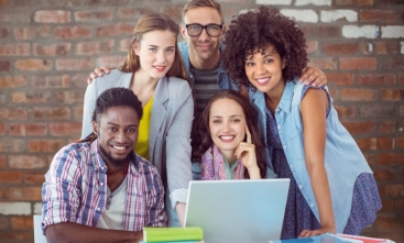 Fotografia ilustrativa de cinco pessoas sorrindo para a foto, três mulheres e dois homens. À frente deles existem livros e um notebook.