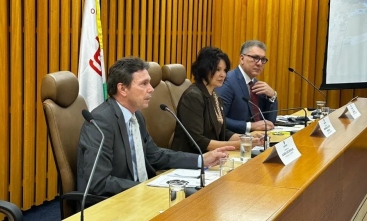 Foto registrada em ângulo de 45 graus mostra dois homens de terno e uma mulher sentados à mesa de um evento