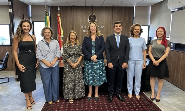 Seis mulheres vestidas formalmente e um homem de terno escuro posam de pé para a foto, sorridentes