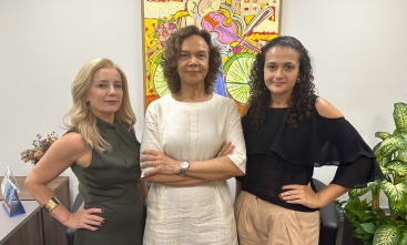 Três mulheres vestidas formalmente em pé, em pose desafiadora, diante de um quadro colorido