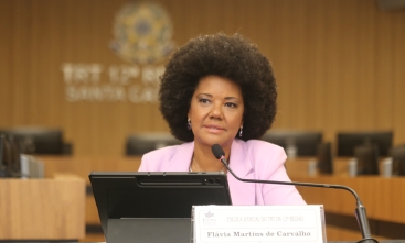 Mulher negra, de cabelo crespo, estilo "black power", fala ao microfone sentada em uma mesa de evento