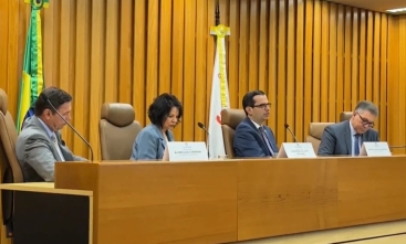 Três homens de terno e uma mulher vestida formalmente sentados em uma mesa de eventos, em tom madeira-ouro, com uma bandeira do Brasil atrás