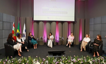 Grupo de sete mulheres sentadas à frente de um auditório, sobre um tablado, formando um semicírculo. Uma delas fala ao microfone